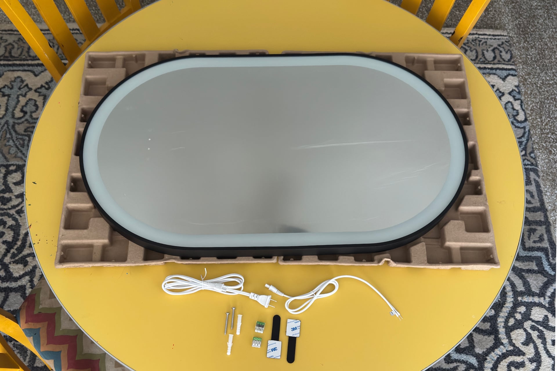 Picture of the smart mirror laying on a table, with its cables and included mounting accessories placed at the bottom of the image.