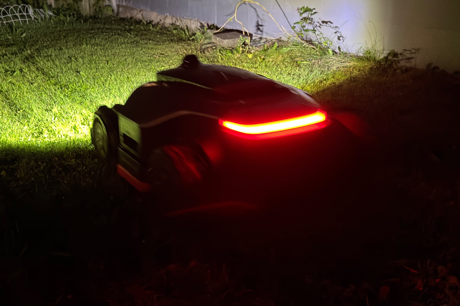 Picture of the Dreame A3 Awd Pro 3500 mowing at night. Its rear red LED strip is clearly visible, while ahead, its LED headlight brightly lights up the yard.