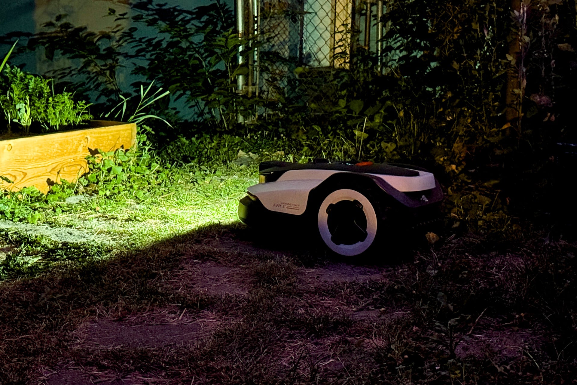 Picture of the Roborock Rockneo Q110H mowing at night, its flashlight lighting up a garden bed in front of it.